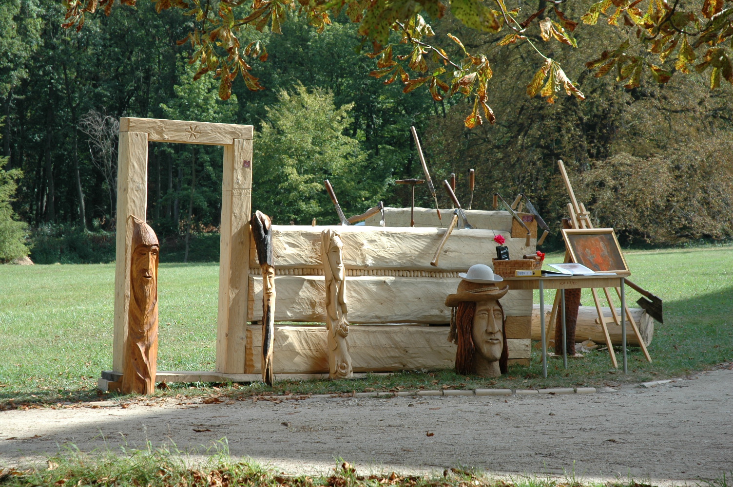 Ręcznie rzeźbione drewniane elementy architektoniczne i dekoracyjne, w tym rama drzwiowa i rzeźby twarzy, wystawione na zewnątrz z narzędziami rzeźbiarskimi i sztalugą w tle.